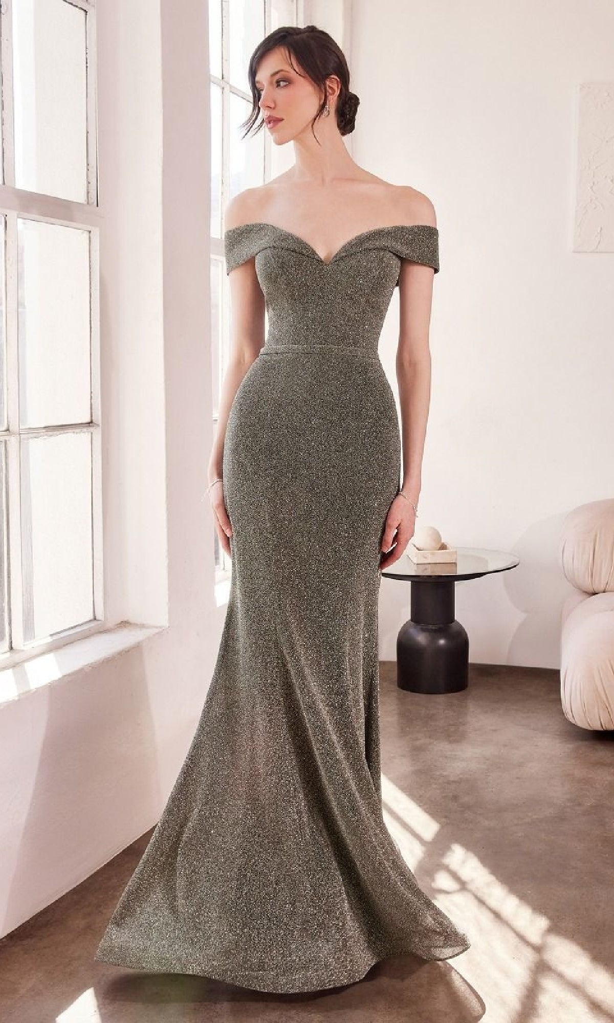 A woman stands indoors in the Off-Shoulder Sweetheart Prom Gown: Amoris T602, a floor-length, fitted evening dress with shimmering glitter details, as her hair is styled in a chic low updo.
